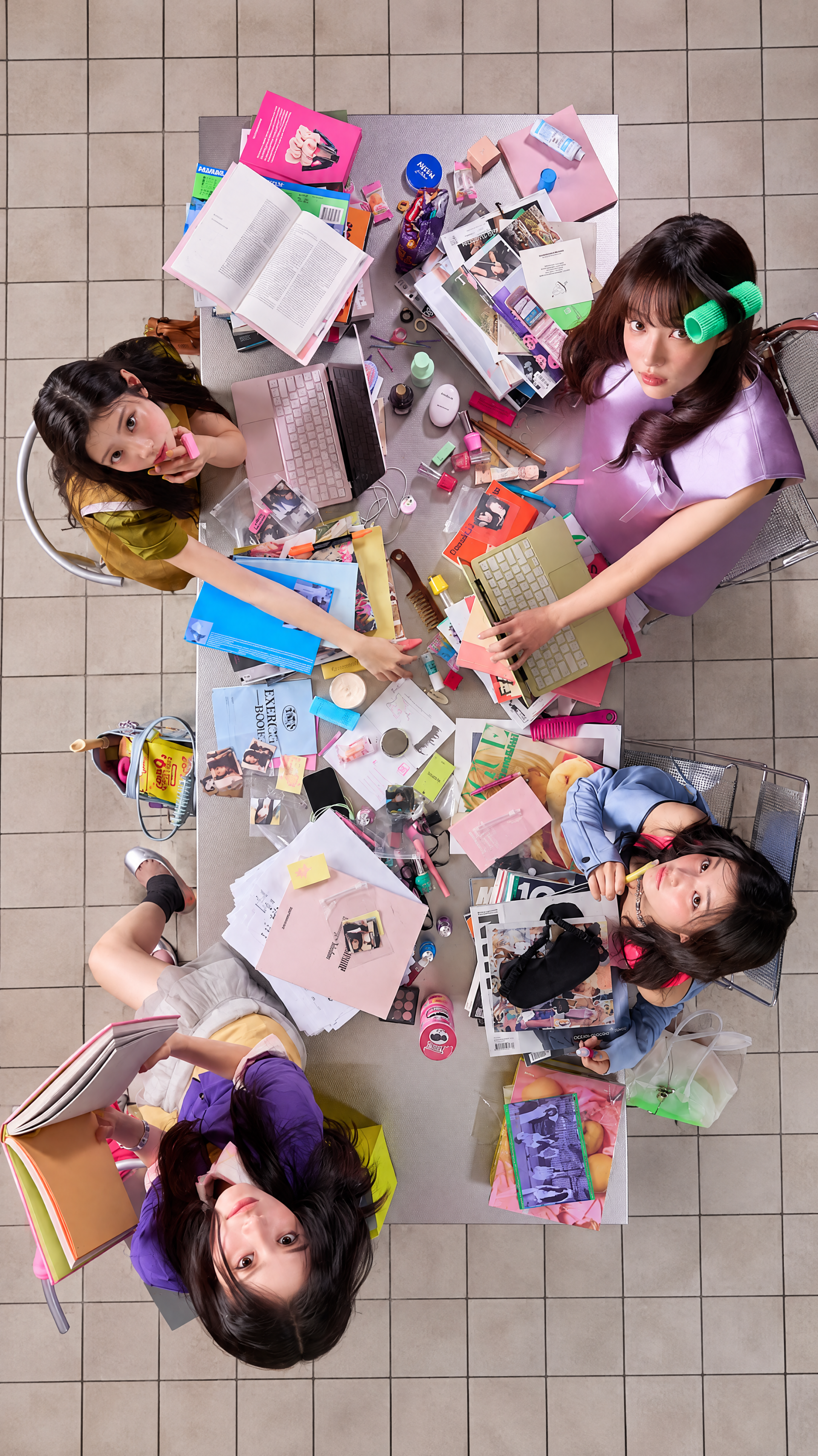 ILLIT break even iPhone wallpaper featuring women studying around a cluttered desk in a colorful overhead scene.