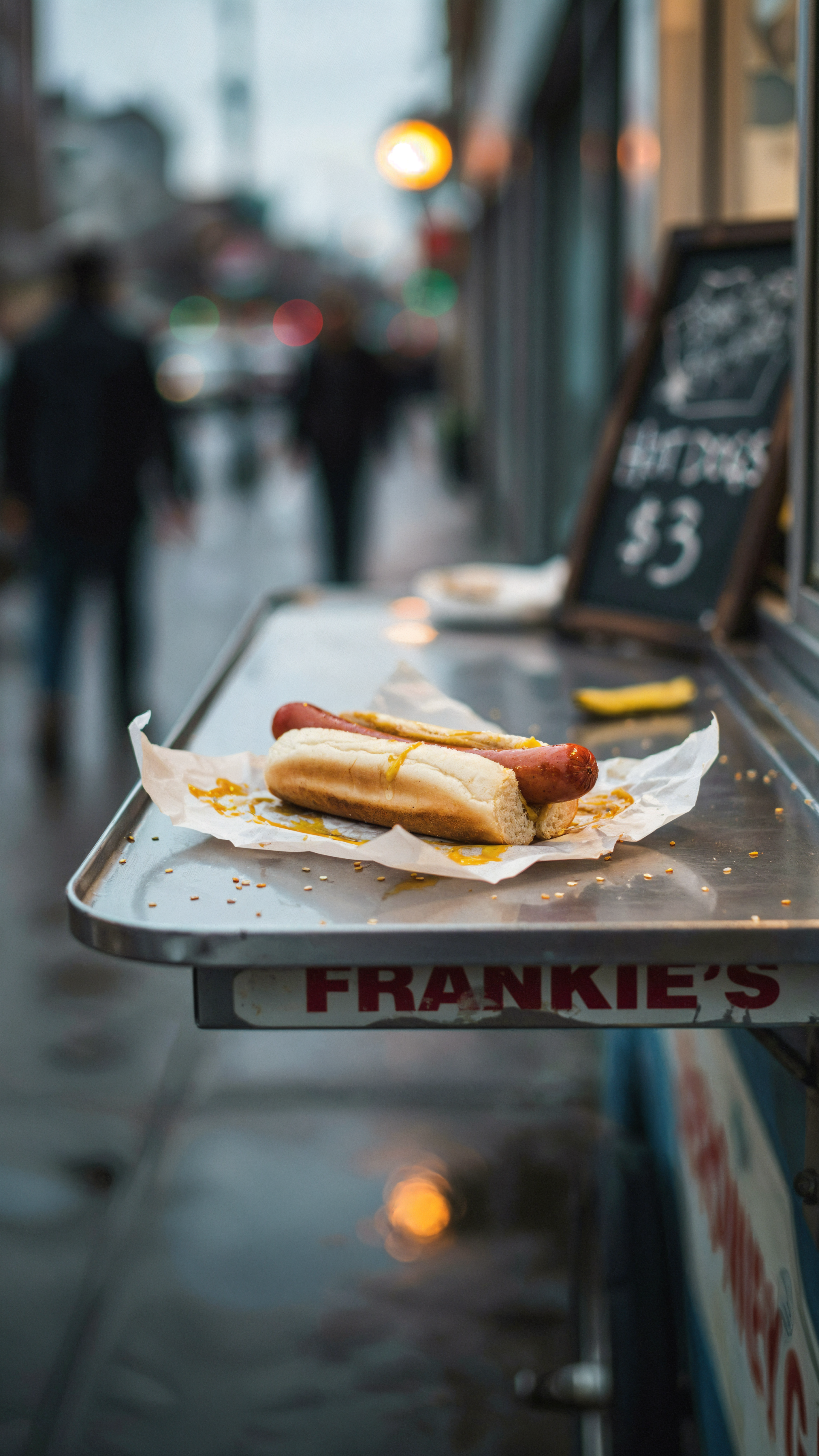 A hot dog on a paper tray at a busy street food stand, shown in a vertical iPhone wallpaper.