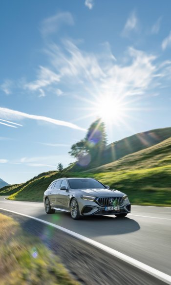 A sleek Mercedes-AMG E 53 Hybrid driving along a scenic road under a bright sky, featuring lush green hills and a radiant sun, makes for a striking phone wallpaper.