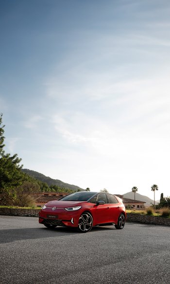 A striking red Volkswagen ID.3 GTX parked on a scenic road, surrounded by greenery and buildings, makes an impressive phone wallpaper.