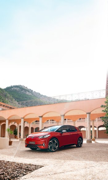 A vibrant red Volkswagen ID.3 GTX parked in a scenic courtyard with arches and mountains in the background, designed as an eye-catching phone wallpaper.