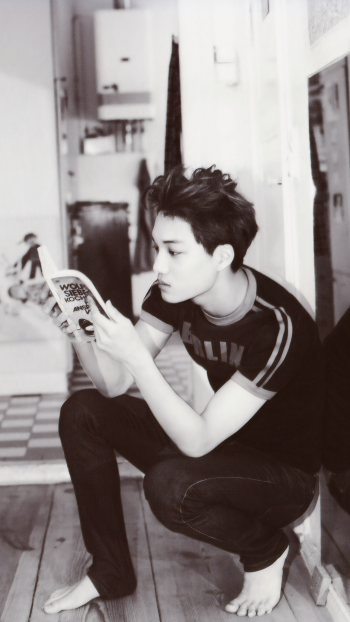 Phone wallpaper of EXO's Kai from the MAMA photoshoot, captured in a black-and-white candid moment reading a book indoors, compatible with iPhones and Android phones.