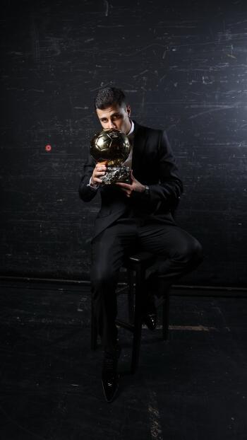 Rodri Hernández, dressed in a black suit, sits on a stool holding the prestigious Ballon d'Or trophy against a dark background, capturing the essence of soccer excellence.