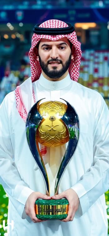 A proud moment featuring a man in traditional attire holding a championship trophy, celebrating Al Hilal SFC's achievement in a vibrant sports arena.