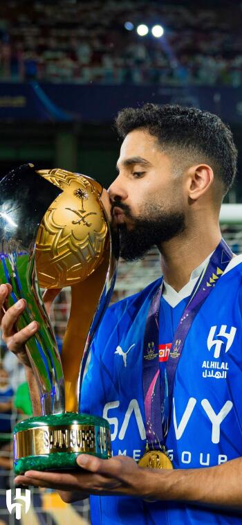 A player from Al Hilal SFC kisses their trophy, celebrating a victory. The athlete wears a blue jersey and a medal, capturing a triumphant moment in sports.