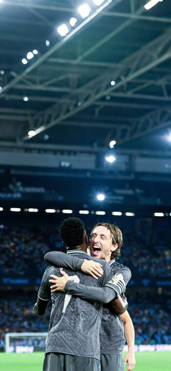 Luka Modrić celebrates passionately with a teammate during a Real Madrid C.F. match, surrounded by an energetic crowd under bright stadium lights. A striking soccer phone wallpaper.