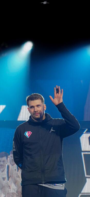 Luka Dončić waves from the stage, illuminated by dramatic lights, capturing the essence of basketball and the excitement of the NBA in this dynamic phone wallpaper.