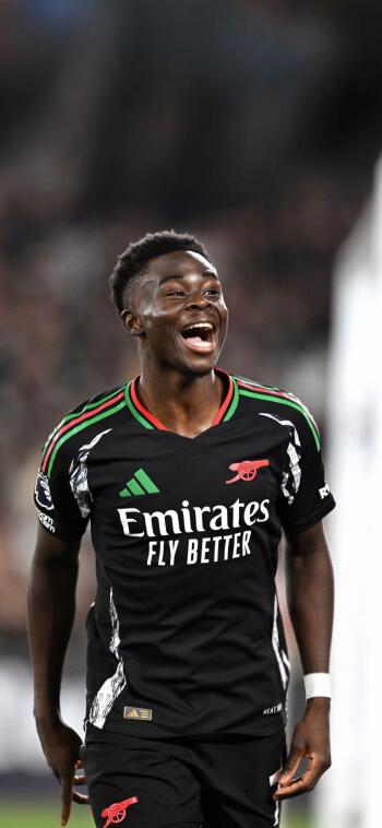 Bukayo Saka celebrates joyfully while wearing Arsenal F.C.'s black away kit, showcasing his enthusiasm for soccer in this dynamic phone wallpaper.