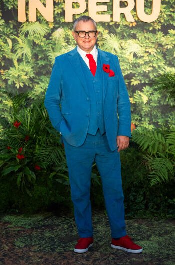 Hugh Bonneville poses confidently in a vibrant blue suit with a red tie and flower, surrounded by lush greens, promoting the movie Paddington in Peru.