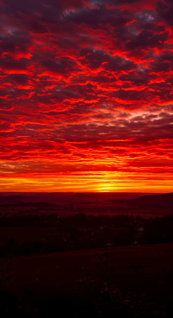 A breathtaking dawn scene with vibrant red and orange clouds illuminating the sky as the sun rises over the horizon, creating a stunning phone wallpaper.