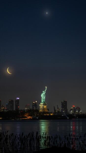 Night view of the illuminated Statue of Liberty in New York with a crescent moon, captured as a vertical phone wallpaper compatible with iPhones and Android devices.