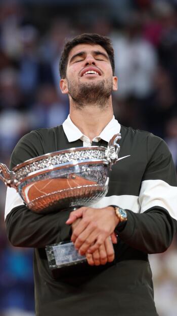 Phone wallpaper of tennis player Carlos Alcaraz celebrating with the French Open (Roland Garros) trophy, captured in a sports moment. Compatible with iPhones and Android phones.