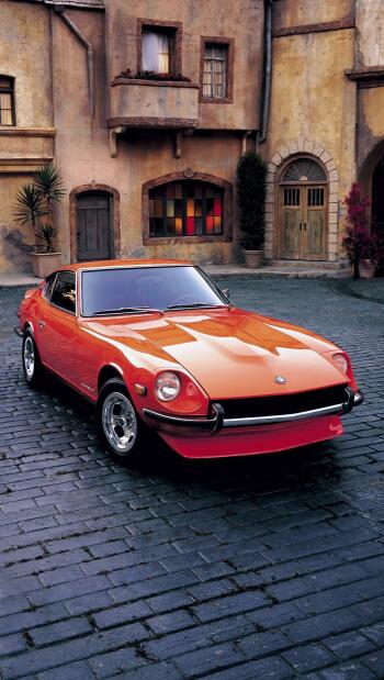 Orange vintage Datsun 240Z (Nissan) parked on a cobblestone street before an old stucco building — vertical phone wallpaper formatted for iPhone and Android.