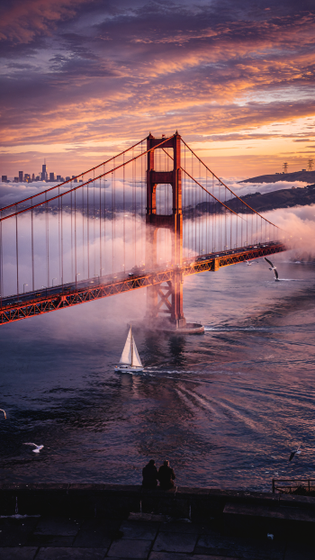 Vertical phone wallpaper of the Golden Gate Bridge at sunrise: fog spilling under the span, a sailboat below and two silhouetted figures on the shore.