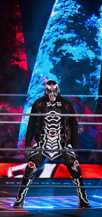 Phone wallpaper showing a masked WWE wrestler in black skeletal ring gear standing in the ring under dramatic blue and red arena lighting, sports action vibe.