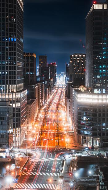 Vertical phone wallpaper of a Japanese cityscape at night: illuminated downtown avenue lined with towering buildings, long light trails and a glowing street grid.