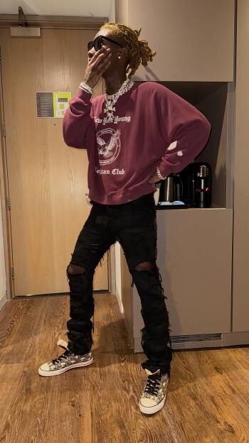 Phone wallpaper of a rapper with blonde dreadlocks and sunglasses, wearing a burgundy sweatshirt, heavy chains and ripped black jeans, striking a casual pose in an indoor hallway.
