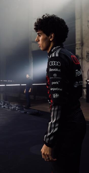 Phone wallpaper: profile of a young man in an Audi F1 racing suit standing in a dim garage, dramatic side lighting and moody atmosphere.