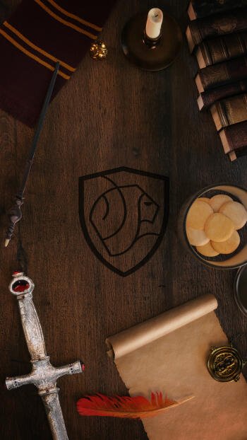Phone wallpaper: Harry Potter Gryffindor scene — wooden desk with etched lion crest, sword, red-and-gold scarf, candle, stacked books, parchment, quill and coins.