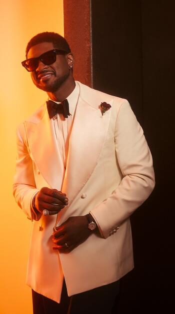 Phone wallpaper of a man in a white tuxedo and sunglasses, styled with rapper flair at an Oscars-themed red carpet, posing like an event usher against warm orange light.