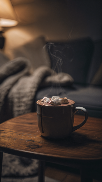 iPhone wallpaper showing a steaming mug of hot cocoa topped with marshmallows on a wooden table, warm lamp glow and a blurred cozy blanket and sofa in the background.