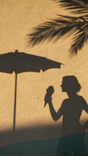 iPhone wallpaper: summer shadow silhouette of a person holding an ice cream beneath a beach umbrella, palm frond shadows cast on a warm sandy wall.