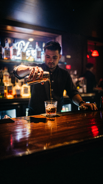 iPhone wallpaper: bartender pouring whiskey into a glass at a dim, neon-lit bar, warm reflections on a polished wooden counter.