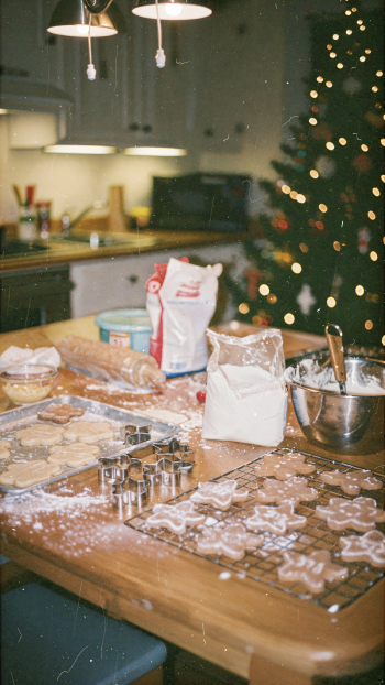 iPhone wallpaper of cozy holiday baking: cookie cutters, dough, flour and a mixing bowl on a kitchen table, with warm, blurred Christmas tree lights in the background.