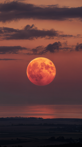Vertical iPhone wallpaper of a large crimson moon rising over a dark coastline, warm orange-red sky and scattered clouds reflected on a calm sea.