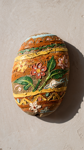 Portrait iPhone wallpaper of a painted rock decorated with colorful floral and geometric bands in warm earth tones, casting a soft shadow on a pale textured wall.