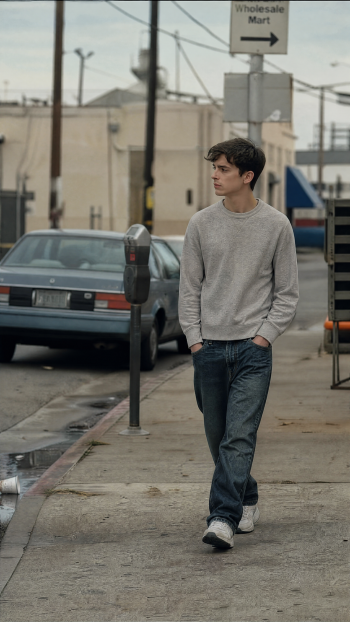 Normcore iPhone wallpaper: person in a grey sweatshirt and jeans walking along a worn urban sidewalk past parked cars and a Wholesale Mart sign.