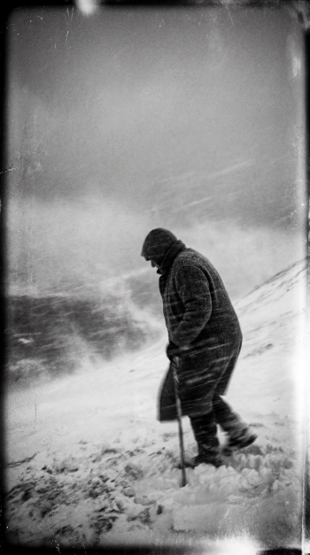 Black-and-white iPhone wallpaper showing a lone figure trudging through a blizzard, hunched against the wind with a walking stick, snow and grainy texture filling a stark, desolate landscape.