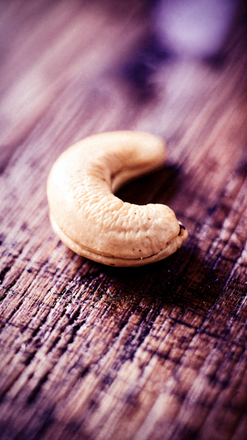Close-up of a single cashew on weathered wood with shallow depth of field, presented in a vertical iPhone wallpaper crop.