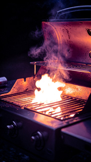 iPhone wallpaper showing a close-up grill with bright flames and curling smoke over hot metal grates, warm orange glow against a dark background.