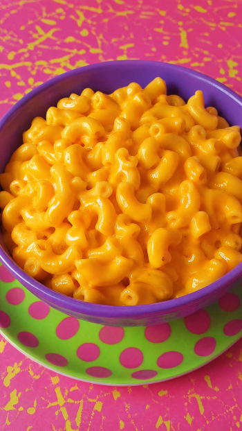 iPhone wallpaper showing creamy mac and cheese in a purple bowl on a green polka-dot plate against a bright pink textured background