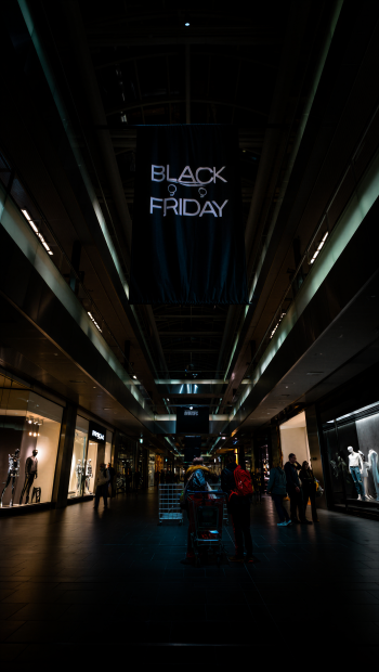Phone wallpaper: dim mall interior at night with illuminated BLACK FRIDAY banner overhead, silhouetted shoppers and a cart centered between lit store windows.