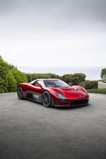 Portrait iPhone wallpaper of a red McLaren McL 6GT sports car parked on a coastal driveway, framed by greenery and sea in the background.