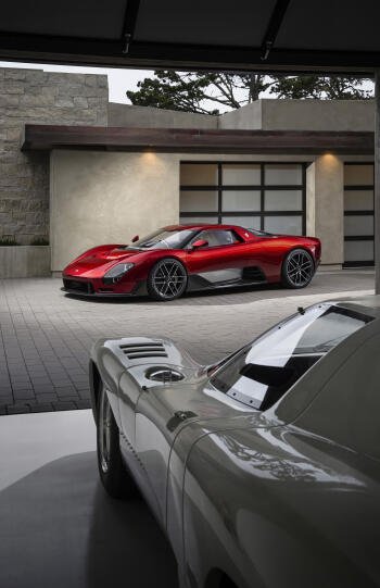 Vertical iPhone wallpaper of a red McLaren McL 6GT supercar parked by a modern garage, another classic sports car vehicle in soft-focus foreground.