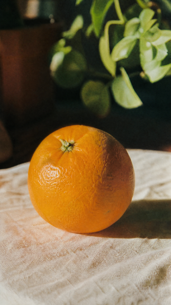 Phone wallpaper portrait: close-up of a bright orange fruit on a white cloth, warm orange color tones with green leaves and a lemon‑citrus accent in the background.