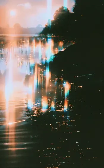 Vertical iPhone wallpaper of a marshy lakeshore at dusk: silhouetted trees and reeds with warm orange and cool teal light streaks reflecting along rippling water.
