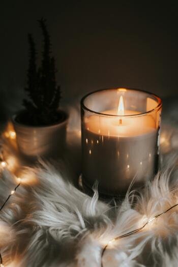 iPhone wallpaper of a lit candle in a glass jar on faux fur with fairy lights and a potted plant, radiating a cozy Kitchen Witch aesthetic.