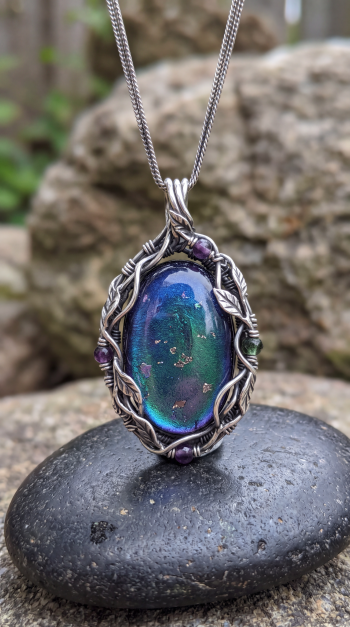 Phone wallpaper showing a close-up wire-wrapped gemstone pendant necklace: iridescent blue-green cabochon set in silver leaf and vine wirework resting on a pebble.