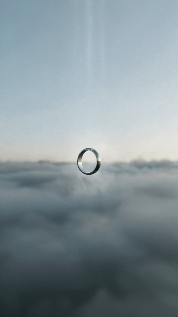 iPhone wallpaper showing a wedding ring suspended like a droplet above misty clouds under a pale sky with a faint vertical beam of light.