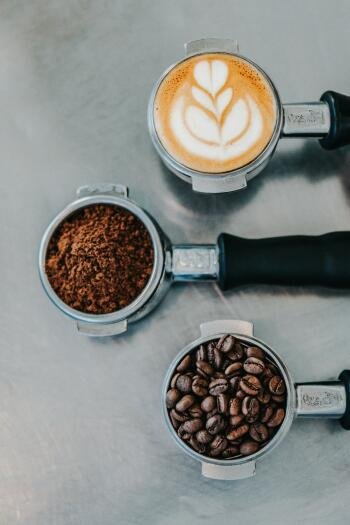 Phone wallpaper Espresso Fall: three espresso portafilters on a gray surface — a latte with leaf latte art, ground coffee, and whole beans; cozy coffee mood.
