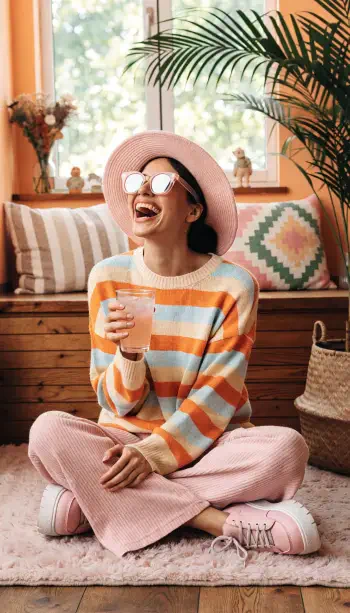 Joyful woman in sunglasses and a sun hat sits cross-legged holding a drink; striped sweater and pastel room create a bright iPhone wallpaper.