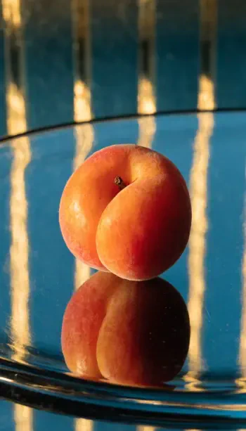 Still-life iPhone wallpaper of a ripe apricot/peach fruit on a reflective surface, warm light casting vertical stripe reflections.