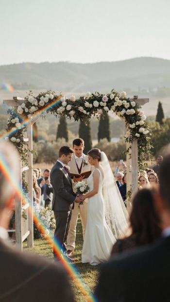 Bride and groom exchange vows beneath a flower-draped arch at an outdoor wedding marriage ceremony, officiant behind them — vertical iPhone wallpaper photo.