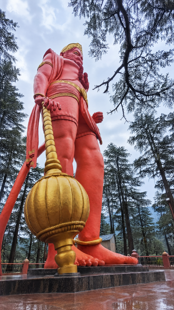 Vertical phone wallpaper of a towering red Lord Hanuman statue holding a golden mace, monument set among pine trees under a cloudy sky.