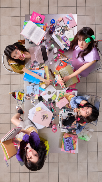 ILLIT break even iPhone wallpaper featuring women studying around a cluttered desk in a colorful overhead scene.