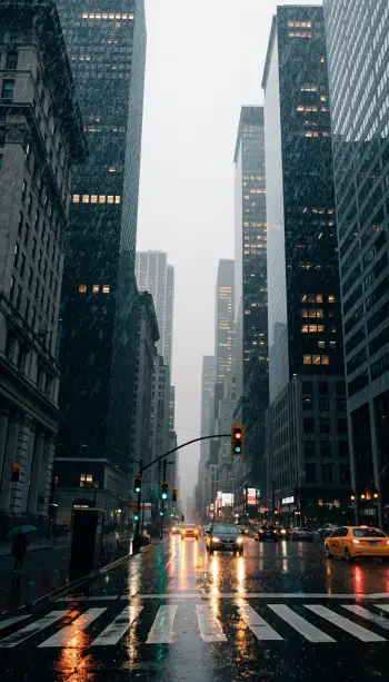 Rainy downtown street lined with illuminated skyscrapers and reflections on wet pavement, vertical iPhone wallpaper.